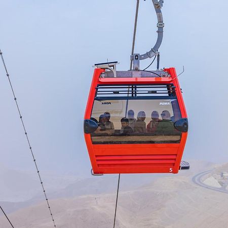 โทลิป รีสอร์ต เอล กาลาลา มาเจสติก Hotel ไอน์ ซกนา ภายนอก รูปภาพ The photo shows a red gondola lift suspended in the air, traveling along cables. Inside the gondola, a group of people can be seen. The background features a hazy landscape, possibly mountains or hills, indicating an elevated area. The scene conveys 