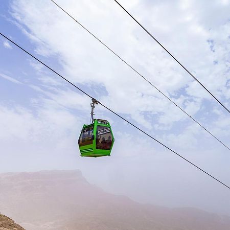 โทลิป รีสอร์ต เอล กาลาลา มาเจสติก Hotel ไอน์ ซกนา ภายนอก รูปภาพ The photo shows a green cable car suspended in the air, traveling along overhead cables. The backdrop features a hazy landscape, likely mountainous or elevated terrain, with a cloudy sky. The scene suggests a scenic transportation method, possibly us